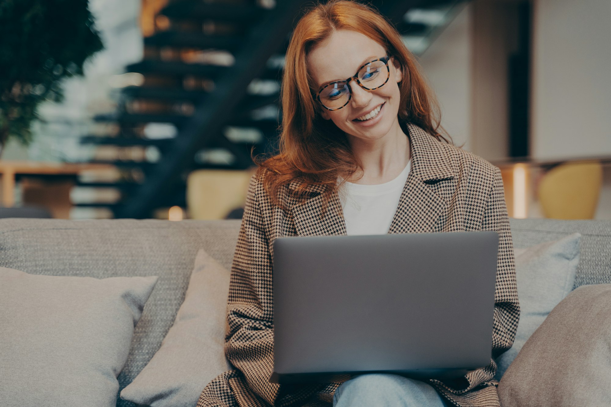 Happy female office worker chatting on laptop while sitting on grey couch at office